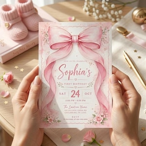 Peut inclure: Une invitation rose pour le premier anniversaire de Sophia, le 24 octobre. La carte présente un grand nœud rose et un motif de ruban, avec des accents floraux et les détails de l'événement. La carte est tenue par une personne.