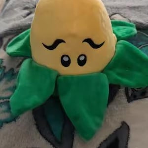 May include: A plush toy in the shape of a sunflower with a yellow head, green petals, and a smiling face. The toy is designed to resemble a character from the Plants vs. Zombies game series.
