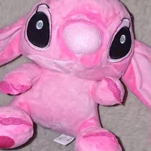 Puede incluir: Un peluche rosa de un personaje de dibujos animados con grandes ojos negros, una nariz rosa y orejas grandes. El juguete tiene una peque&ntilde;a corona rosa en la cabeza y est&aacute; hecho de material suave y difuso. El juguete est&aacute; sobre una superficie gris y difusa.