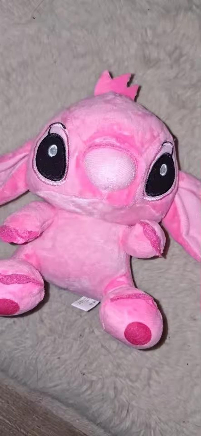 Puede incluir: Un peluche rosa de un personaje de dibujos animados con grandes ojos negros, una nariz rosa y orejas grandes. El juguete tiene una peque&ntilde;a corona rosa en la cabeza y est&aacute; hecho de material suave y difuso. El juguete est&aacute; sobre una superficie gris y difusa.