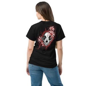 Puede incluir: Una camiseta negra de manga corta con un estampado gráfico en la espalda. El diseño muestra un cráneo animal blanco detallado rodeado de elementos florales rojos y líneas curvas, creando un contraste marcado sobre la tela oscura.