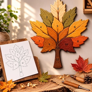 May include: A wooden wall art piece shaped like a large maple leaf, composed of twelve numbered segments in shades of cream, green, yellow, orange, and brown. A matching paper template with numbered outlines is displayed on a wooden stand nearby.