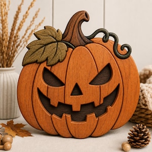 Puede incluir: Una decoración de madera tallada con forma de calabaza que presenta una cara de jack-o'-lantern con ojos triangulares y una amplia sonrisa dentada. La calabaza es naranja con un tallo marrón oscuro, una hoja verde y un zarcillo marrón oscuro.