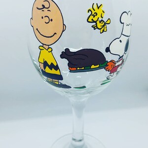 Pode incluir: Uma ta&ccedil;a de vinho de vidro transparente com um desenho pintado com personagens da tira de quadrinhos Peanuts. Os personagens incluem Charlie Brown, Snoopy e Woodstock. O desenho tamb&eacute;m inclui um peru de A&ccedil;&atilde;o de Gra&ccedil;as.