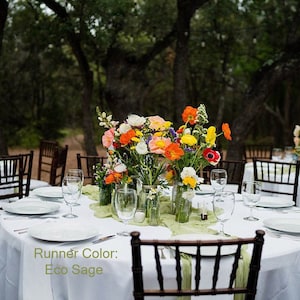 May include: A white tablecloth with a green runner, set with a floral centerpiece in mason jars. The centerpiece features a variety of flowers in shades of orange, yellow, pink, and white. The table is set with clear glass plates, silverware, and wine glasses. The background is a blurred image of trees.