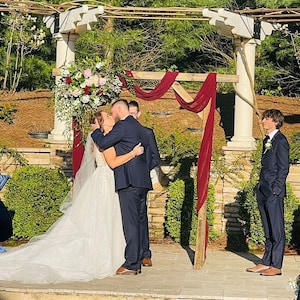 May include: A wedding ceremony taking place outdoors under a wooden archway decorated with burgundy fabric and greenery. The couple is kissing, and a guest is standing to the right of the archway.