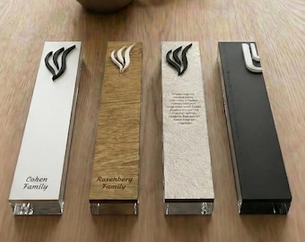 May include: Four decorative mezuzah cases in white, wood grain, textured, and black. Each case has a silver-colored emblem at the top and a clear base. The white case reads "Cohen Family". The wood grain case reads "Rosenberg Family".