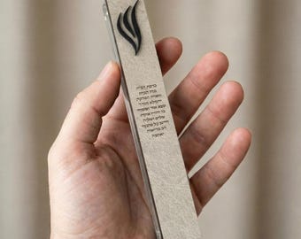 May include: A rectangular mezuzah case with a textured, light beige surface. The case features a black, stylized flame design at the top and Hebrew text. The mezuzah is held in a hand against a neutral background.