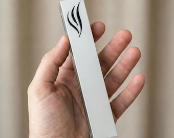 May include: A silver-colored mezuzah case with a black flame design. The rectangular case is held in a hand, showcasing its polished surface and minimalist aesthetic. The design is a modern take on a traditional religious item.