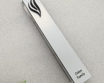 May include: A rectangular, silver-colored mezuzah case with a black flame design at the top. The text "Cohen Family" is printed in black at the bottom. The case is displayed on a white surface.