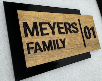 May include: A rectangular wooden sign with the words "MEYERS FAMILY" in black, bold letters. The number "01" is on the right side. The sign has a wood grain pattern and is set against a black background.