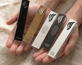May include: Five rectangular mezuzah cases in various colors and textures, including black, wood grain, and white. Each case features a decorative emblem and family name. The cases are held in open hands.
