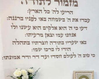 May include: A wall decal with Hebrew text in a stylish font. The text is in a light brown color and is arranged in multiple lines. A vase of white roses is visible at the bottom left.