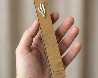 May include: A wooden mezuzah case with a light brown finish, featuring a white flame design. The text "Rosenberg Family" is engraved on the bottom. The mezuzah is held in a hand.