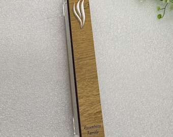 May include: A rectangular wooden mezuzah case with a light brown wood grain finish. It features a silver-colored, stylized flame design and the engraved text "Rosenberg Family" at the bottom. The case has a clear acrylic side.