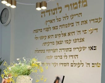 May include: A wall with gold Hebrew text, a clock, and a modern light fixture. A vase of flowers sits on a table in front of the wall. The text is a religious passage.