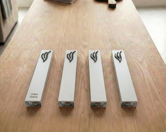 May include: Four white mezuzah cases with a black decorative design on top, arranged on a wooden table. The bottom of each case is clear. One case has the text "Cohen Family".