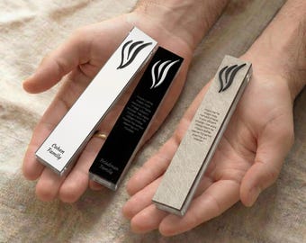 May include: Three rectangular mezuzah cases in white, black, and beige are displayed. Each case features a stylized flame design and text. The white case is labeled "Cohen Family," the black case "Friedman Family."