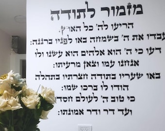 May include: A white wall with black Hebrew text. The text is a religious passage, likely a prayer or psalm. A bouquet of white and yellow flowers sits in front of the wall. A power outlet is visible.