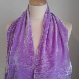 Puede incluir: Bufanda de terciopelo lavanda drapeada sobre un maniquí. La bufanda tiene un aspecto suave y texturizado con sutiles variaciones de color. La tela está fruncida y drapeada para crear un aspecto fluido y elegante. La bufanda es de color sólido.