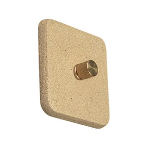 Pode incluir: Uma placa de interruptor de parede quadrada bege claro com uma superfície texturizada. Um pequeno botão cilíndrico dourado está centrado na placa. O botão tem um padrão texturizado e canelado.