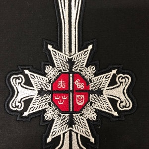 Puede incluir: Un parche bordado blanco con un centro rojo. El centro tiene cuatro secciones con símbolos y los números "666". El parche tiene la forma de una cruz con una parte superior larga y puntiaguda.