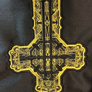 May include: A gold embroidered patch in the shape of an inverted cross. The cross is detailed with intricate patterns and designs.