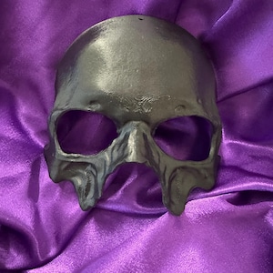 Può includere: Una maschera a teschio grigio scuro con grandi orbite oculari e dettagliati lineamenti del viso. La maschera è realizzata in un materiale solido ed è esposta su uno sfondo viola strutturato.