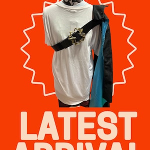 May include: A white t-shirt with a black belt and a blue and black cape. The belt has a large gold buckle with a decorative design. The image is set against a bright orange background with the text "NEW! NEW! NEW! NEW! NEW! NEW!" at the top and "LATEST ARRIVAL" at the bottom.