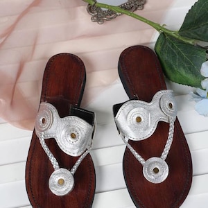 Könnte beinhalten: Ein Paar braune Ledersandalen mit silbernen Verzierungen. Die Sandalen haben silberne Riemen mit kreisförmigen Akzenten und einen geflochtenen Zehenriemen. Die Sohlen sind dunkelbraun. Die Sandalen sind neu.