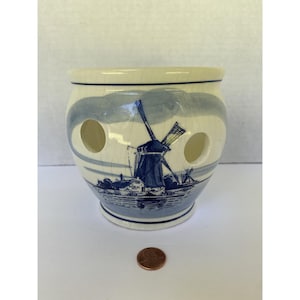 May include: A ceramic onion keeper with a blue and white windmill design. The keeper has three circular holes and a crackled finish. The windmill scene is depicted on the front, with a landscape at the base. A penny is next to the keeper.
