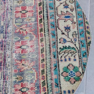 Könnte beinhalten: Ausschnitt eines runden, Vintage-Teppichs mit verblasstem, abgenutztem Aussehen. Der Teppich weist eine Mischung aus Farben auf, darunter Rosa, Blau, Creme und Hauch von Grün. Das Design umfasst florale und geometrische Muster mit einem türkisfarbenen und schwarzen Rand.