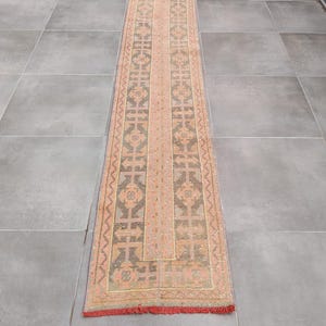 Puede incluir: Una alfombra larga y rectangular con un patrón geométrico central en tonos melocotón, gris y marrón. La alfombra tiene un borde con flecos y un diseño repetido a lo largo de los bordes. La alfombra es probablemente de material tejido.