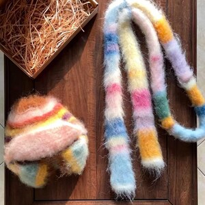 Peut inclure: Un ensemble chapeau et écharpe duveteux aux couleurs pastel de rose, jaune, bleu et orange. Le chapeau est rond avec un bord, et l'écharpe est longue et rectangulaire. Les articles sont présentés sur une surface en bois foncé.
