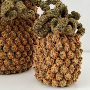May include: Two handmade crochet bags in shades of brown and green. The bags have a textured, bumpy surface and a decorative, looped top. The yarn appears to be a blend of colors, giving the bags a rustic, natural look.