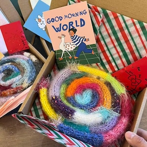 Pode incluir: Uma caixa de cartão contém um artigo de malha em espiral colorido, um cartão de felicitações com o texto "GOOD MORNING WORLD" e outros cartões pequenos. A espiral é uma mistura de cores brilhantes, incluindo amarelo, laranja, azul e rosa.