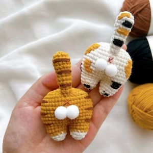May include: Two handmade crochet cat butt ornaments. One is yellow with brown stripes, the other is white with orange and black spots. Both have white pom-pom details and are held in a hand.