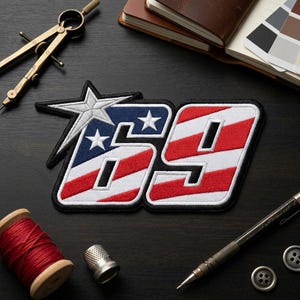 May include: An embroidered patch featuring the number 69 in a bold, block font. The design incorporates a red and white striped pattern with a blue section containing two white stars. A large, silver-toned star is attached to the upper left corner.