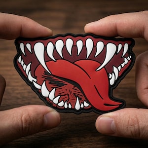 May include: A patch featuring a stylized open mouth with sharp white teeth, a red tongue, and a maroon interior. The patch has a black border and is held between two hands.