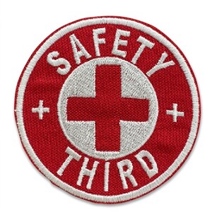 May include: A circular embroidered patch featuring a red background with a white border. In the center is a white cross. The text SAFETY is arched above the cross and THIRD is arched below, both in white capital letters. Two small white crosses are on the sides.