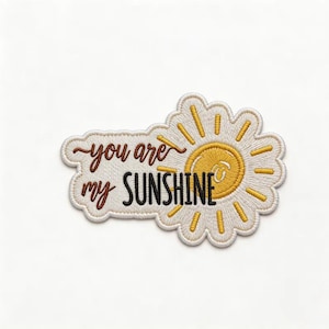 May include: An embroidered patch featuring a white daisy-shaped border with a yellow sun center. The text "you are my SUNSHINE" is stitched in brown and black thread across the white background, with the word SUNSHINE in bold black letters.