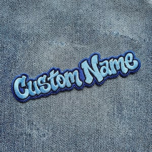 May include: A blue embroidered patch with the words "Custom Name" in a bubble font. The patch has a light blue outline and is set against a denim background.