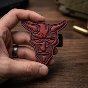 May include: A red devil face patch with a detailed design, featuring horns, a stern expression, and a goatee. The patch is held against a blurred background, showcasing its intricate features and dark red color.