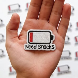 May include: An embroidered patch featuring a white battery icon with a red low-battery indicator and a black border. Below the battery icon, the text "Need Snacks" is embroidered in black thread against a white background with a black border.