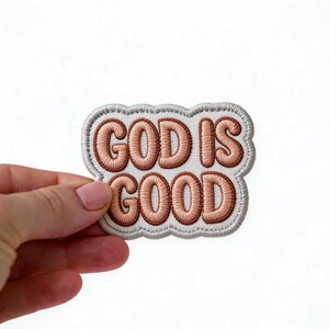 May include: An embroidered patch with a white background and a scalloped border in light gray. The text GOD IS GOOD is stitched in a rounded, bubble-style font using a soft peach-colored thread, creating a textured and raised appearance.