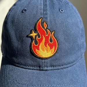 May include: A navy blue baseball cap with a vibrant embroidered patch of a flame design. The flame features shades of red, orange, and yellow, with a small star accent. The cap has a curved brim and a classic six-panel design.