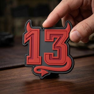 May include: A black and red patch featuring the number 13 with devil horns and a tail. The number is red with a black outline and sits on a gray background. The patch is held by a hand.