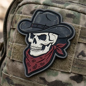 May include: A black and white skull patch wearing a cowboy hat and a red bandana with a paisley pattern. The patch is attached to a camouflage background.