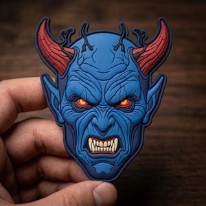 May include: A blue devil face patch with red horns and glowing orange eyes. The patch features a detailed design with sharp teeth and a menacing expression. The patch is held in a hand against a wooden background.