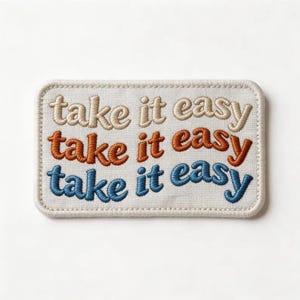 May include: A rectangular embroidered patch on a light cream fabric base. The patch features the text "take it easy" stitched in three horizontal rows, with the top row in beige, the middle row in burnt orange, and the bottom row in blue.